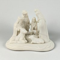 A monochromatic white porcelain nativity scene featuring a smooth, matte texture. The sculpture depicts a group of figures and animals on a rounded base, including the Holy Family and Wise Men, characterized by soft silhouettes and refined, uniform coloring.