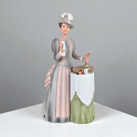 Matte porcelain figurine of a Victorian woman in a grey and pink striped bustle dress. She wears a feathered hat and stands beside a small draped table holding a vanity case and perfume bottles. The palette features soft sage green, blush, and slate grey.