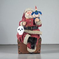 A fabric-clothed Santa figurine sits atop a wooden toy chest, holding a small wooden soldier puppet. A miniature white skull rests on his knee. He wears a red suit with popcorn-textured trim, a black belt, and shiny black boots.
