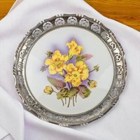 A circular porcelain plate with a wide, pierced silver-toned metal rim featuring a repeating rose pattern. The center of the plate is decorated with a hand-painted cluster of bright yellow flowers and green leaves against a soft purple and white gradient background.