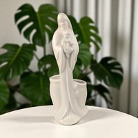 A matte white ceramic figurine of the Madonna holding the infant Jesus. The texture is smooth and non-reflective. The silhouette is tall and fluid, featuring a hollow planter well integrated into the back of the figure.
