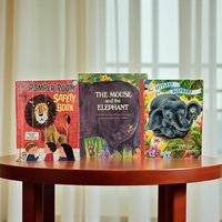 Three vintage children's books stand upright on a polished wood surface. The left cover features a lion and three children in a vibrant mid-century illustration style. The center book shows a large purple elephant silhouette with a small mouse. The right book depicts a blue baby elephant holding a flower.