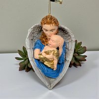 A sculpted resin ornament featuring a red-haired angel in a vibrant blue robe cradling a baby in gold swaddling. The figures are framed by large, textured white wings that form a heart shape at the base.