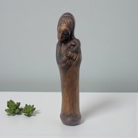 A slender wooden sculpture of the Virgin Mary holding the infant Jesus. The wood features a warm, honey-toned grain and smooth, flowing carved lines that define the figures' robes and serene expressions.