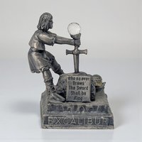 A matte gray pewter figurine of King Arthur pulling the sword Excalibur from a stone. The metal features a weathered, dark patina. A small multifaceted clear crystal sits atop the sword hilt.