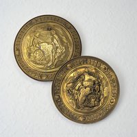 Two circular brass wall plates featuring embossed scenes of a man and woman in 18th-century attire. The metal has a warm golden hue with aged dark patina in the recessed areas and a scalloped rim.