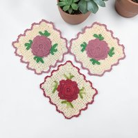 Three circular hand-crocheted pot holders in cream yarn with central dimensional flowers in pink, yellow, and blue. Scalloped edges and integrated hanging loops. Soft textured stitch work with a slight vintage patina.