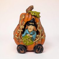 A glossy orange ceramic pumpkin shaped like a carriage. It features a black door, yellow glowing windows, and four black wheels. A small black stem sits on top with curly green vines trailing down the sides.