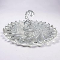 A clear circular glass serving tray with a prominent center handle. It features a dramatic swirling ribbed texture radiating from the center to the scalloped edge. The glass is thick, transparent, and highly reflective.