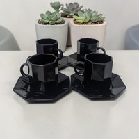 Four small black glass coffee cups and matching saucers. The pieces feature a distinct octagonal geometric shape with a high-shine, opaque black finish. The cups have a small side handle and flat angular sides.
