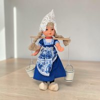 A plastic doll wearing a traditional Dutch costume including a pointed white lace cap and blue striped skirt. She carries a wooden yoke across her shoulders with two small blue and white ceramic pails hanging from metal chains.