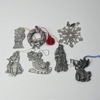 A collection of seven vintage metal ornaments. The set includes detailed pewter figures of Santa in various poses, an angel with a horn, a red-accented wreath with a tassel, and a delicate silver-plated snowflake with an openwork design.