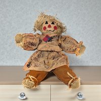 A tall, handmade scarecrow figure wearing a plaid flannel shirt, denim overalls, and a pointed burlap hat. The face is crafted from fabric with hand-drawn features, and straw-like raffia tufts peek out from the sleeves and hat.