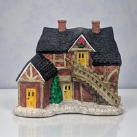 A snow-dusted ceramic village house featuring a steeply pitched roof, handpainted green shutters, and a bright red front door. Small cut-out windows allow light to glow from within the cream-colored structure.
