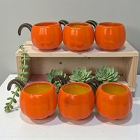 A set of six small ceramic cups shaped like plump orange pumpkins. Each features a green vine-style handle, textured vertical ribbing, and a glossy hand-painted finish.