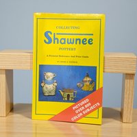 A softcover reference book with a bright yellow cover featuring photographs of various Shawnee pottery pieces, including a teapot and character jars. The text is bold and easy to read, and the binding is clean.