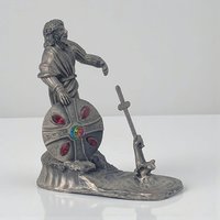 A solid pewter figurine featuring a hand emerging from a swirling pool of water, grasping a detailed sword. The metal has a soft, matte grey finish with intricate textures on the water’s surface and the sword’s hilt.