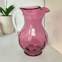 A small cranberry red glass pitcher with a clear, applied handle. The body features a subtle diamond optic pattern that catches the light. The vessel has a wide, flared spout and a bulbous base.