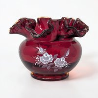 A small, squat glass vase in deep ruby red with a wide, ruffled rim. Delicate white and blue flowers with green leaves are hand-painted on the front. The surface is glossy and smooth with a crimped top edge.