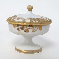 A small, round porcelain lidded box with a creamy white finish. Intricate gold floral patterns and scrolling vine motifs cover the lid and body. The gilding has a slightly raised, textured feel against the smooth porcelain.