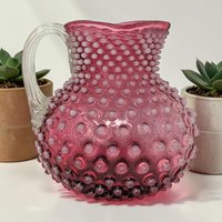 A round glass pitcher in deep cranberry red, covered in a uniform hobnail texture of raised bumps. It features a clear, smooth glass handle and a wide, circular rim. The base is slightly flattened and translucent.