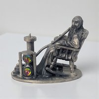 A solid pewter figurine of an elderly bearded man with a long pipe sitting in a rocking chair. A large hourglass stands beside him, containing two iridescent rainbow crystal orbs. The piece has a dark metallic patina and detailed textures.