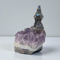 A detailed pewter figurine mounted on a jagged, purple amethyst crystal cluster. The silvery metal features a dark patina in its recessed details, contrasting with the translucent violet hues of the stone base.