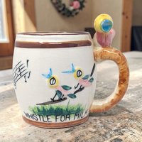 Cream ceramic mug featuring hand-painted whimsical birds on a branch and musical notes. A small sculpted bird sits atop a textured brown handle. Fine crazing visible across the glazed surface with a hand-lettered base.