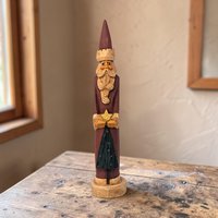 Tall slender hand-carved wooden Santa Claus figurine in a traditional red robe with white trim. The figure holds a small green tree. Natural wood grain visible through the matte paint.