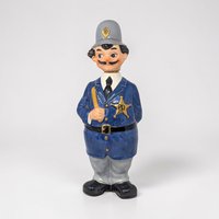 A painted ceramic policeman figurine in a blue uniform and grey helmet with gold "P.D." accents. He has a black mustache, holds a nightstick, and wears a gold star badge. The figure stands on black shoes with hand behind his back.