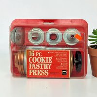 A vintage cookie and pastry press set in its original red and clear plastic blister packaging. The set includes a copper-toned metal barrel with a black turn-handle and various silver-toned metal shaping discs and pastry tips.