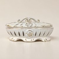 An oval ivory ceramic trinket dish with a matching lid. Both pieces feature vertical reticulated cutouts and gold-painted scroll edges. The lid has a decorative scrolled handle, and the body sits on a scalloped pedestal base with gold accents.