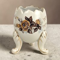 Cream porcelain egg-shaped vase with a jagged rim and three gilded scroll feet. Features a central transfer of golden-brown roses and smaller gold leaf sprigs. Smooth, glazed ceramic texture with gold trim on the feet and scalloped edge.