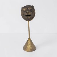A tall brass sculpture featuring a stylized tiger or lion face with large eyes and an open mouth. The mask is mounted on a slender rod fixed to a conical base decorated with sunburst and geometric engravings.