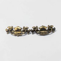 A pair of brass drawer pulls featuring an ornate batwing-shaped backplate and a curved bail handle. The metal has a deep, aged gold patina with visible swirling flourishes and traditional mid-century decorative etching.