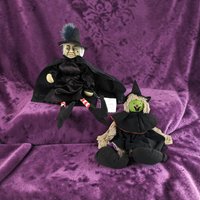 Two witch dolls, one with a realistic sculpted face and feathered hat, the other with a green fabric face and yarn hair. Both wear black cloaks and pointed hats against a purple backdrop.