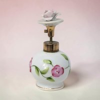 A 3.75-inch bone china perfume bottle with a round bulbous silhouette and a smooth white porcelain texture. It features hand-painted pink roses with green leaves and a sculptural rosebud stopper on a gold-tone metal stem.