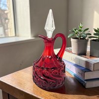 A ruby red glass cruet with a bulbous body featuring a raised, swirling scroll pattern. It has a graceful curved handle and a clear, pointed crystal stopper that sits in the narrow neck.