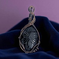 A teardrop-shaped black onyx stone with a deeply carved floral pattern, encased in dense oxidized copper wire weaving. The metal has a dark, antiqued patina with intricate coiled and layered textures.
