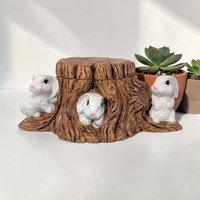 Hand-painted ceramic candy dish shaped like a hollowed-out tree stump. Two white rabbits with pink ears are nestled at the base among green grass. The stump is textured with brown bark and knots.
