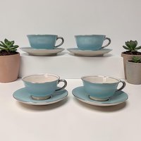 Set of four delicate white fine china tea cups and matching saucers. Each piece features a thin, polished silver rim. The cups have a low, wide profile with a dainty looped handle and a high-gloss finish.