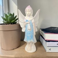 Tall ceramic fairy figurine with a high-gloss finish. She has long flowing hair and large, detailed wings with iridescent accents. She wears a soft pastel gown and stands on a naturalistic base of mushrooms and moss.