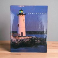A glossy hardcover book featuring a tall white lighthouse on a rocky shore at dusk. The sky and water are deep blue, and the lantern room glows with yellow and green light. The title Lighthouses is printed in white sans-serif capital letters.