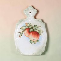 A shield-shaped porcelain wall plaque with a decorative scrolled top and scalloped edges. It features a handpainted center illustration of two red apples on a leafy branch against a pale blue and white background with a soft green border.