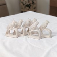 A set of eight matching white porcelain napkin rings. Each ring features a sculpted bird with detailed feather textures, perched on a smooth circular base. The entire piece is finished in a uniform, high-gloss white glaze.