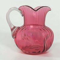 A vibrant cranberry red glass pitcher with a rounded body and a flared, ruffled rim. It features a clear glass handle attached to the side and a polished pontil mark on the bottom. The glass has a rich, translucent glow.