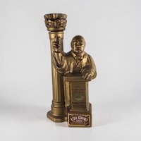 A gold-toned ceramic decanter depicting a bust of Winston Churchill at a podium next to a tall fluted column. The surface has a matte metallic finish with a textured, sculptural silhouette and several original paper labels on the base.