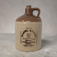 A 7-inch 1974 Daniel Boone bourbon stoneware jug. Features a two-tone cream base and tobacco-brown glazed shoulder with a black graphic of Daniel Boone in a coonskin cap. The cylindrical silhouette includes a finger loop handle and an original cork.