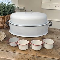 A large oval white enamel roasting pan with a domed lid and black handles. Four small white enamel cups with red rims sit in front. The set shows significant chipping and patina on the enamel surfaces.
