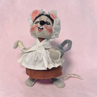 Gray felt mouse with a wide grin and black button nose. She wears a brown felt skirt, a white fabric apron with a cross-over bodice, and a white ruffled mobcap. Her long gray tail is curled behind her against a pink background.